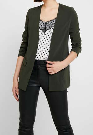 Frau trägt dunkelgrünen Blazer über weißem Top mit Polka-Dots und Spitzenbesatz sowie schwarze Lederhose, hält den Blazer mit einer Hand.
