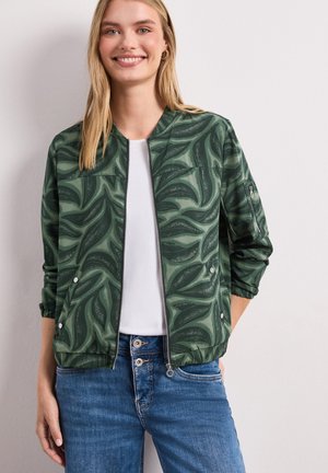 Lächelnde Frau trägt grüne gemusterte Bomberjacke mit Reißverschluss über weißem Shirt und blauen Jeans, steht vor weißer Wand.