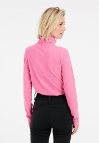 Ružova švelni turtleneck megztinis su ilgemis rankovėmis, prigludusio silueto ir aukštu apykakle, derinamas su juodomis kelnėmis.