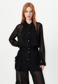 Crna košulja s dugim prozirnim rukavima, s oštrim ovratnikom i srebrnim gumbićima. Košulja je uparena s crnim plisiranim hlačama.