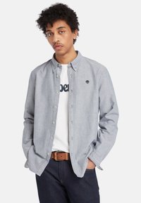 Camisa de botones azul claro con cuello puntiagudo, fabricada en chambray gris, y un pequeño logo en el pecho, llevada sobre una camiseta blanca.
