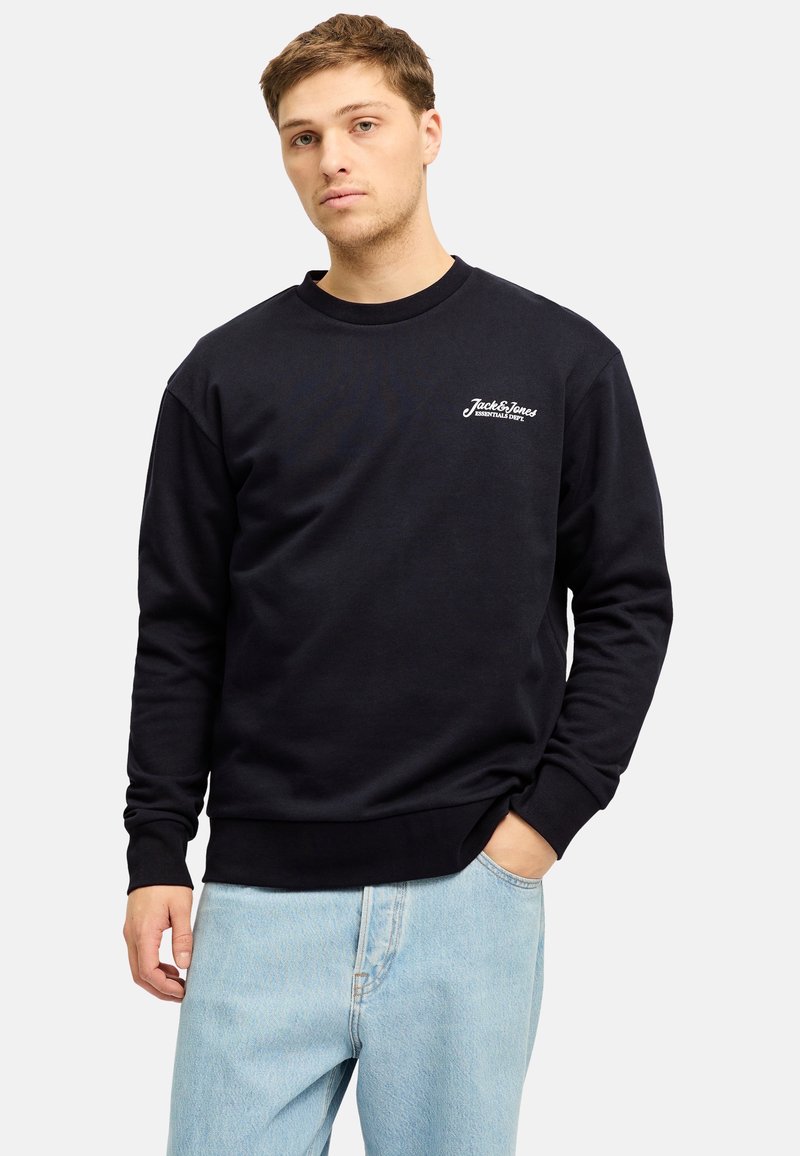 Sudadera negra con cuello y puños de canalé, que presenta un pequeño logo blanco en el pecho. Combinada con jeans de mezclilla azul claro.