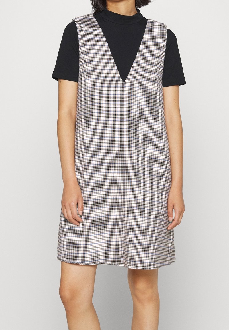 Femme portant une robe sans manches à col en V à carreaux par-dessus une chemise noire à manches courtes sur un fond clair uni.