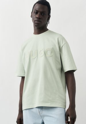 Mann mit dunkler Haut und kurzen Zöpfen trägt ein hellgrünes oversized T-Shirt mit erhabenem "HUGO"-Schriftzug und hellblauen Jeans.