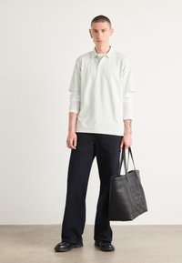 Lichtgroen polo shirt over een wit shirt met lange mouwen, gecombineerd met donkere wijde broek en een zwarte leren tote bag met de ingedrukte tekst "HUGO."