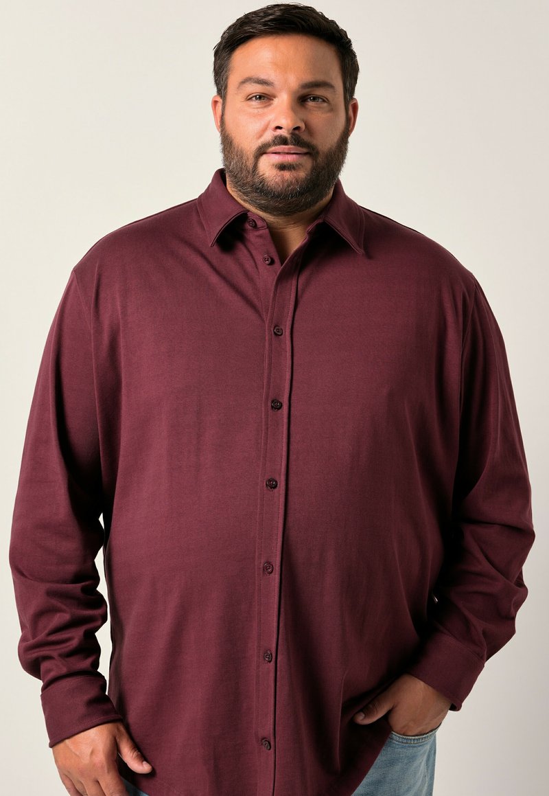 Uomo con barba che indossa una camicia bordeaux a maniche lunghe e jeans azzurri chiari, in piedi con una mano in tasca.