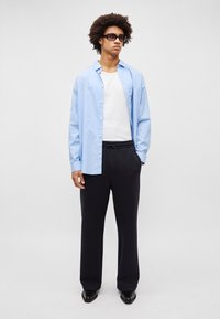 Gestreiftes blaues Hemd über einem weißen T-Shirt, kombiniert mit schwarzen Jogginghosen und schwarzen Schuhen. Das Model hat lockige Haare und eine Sonnenbrille.