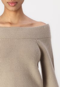 Beige ribbed off-the-shoulder sweater with wide neckline and textured fabric, featuring long sleeves and a fitted silhouette.