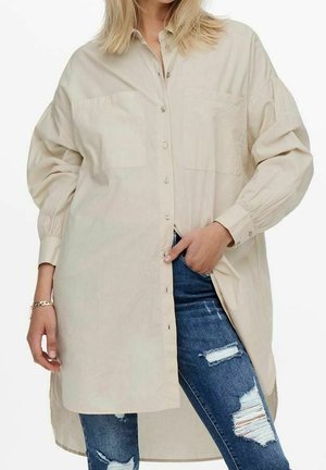 Vaalean beige napituspaita, jossa on kaksi rintataskua, pitkät hihat ja hieman pidempi takaosa, päällä kulutettu sininen farkut.