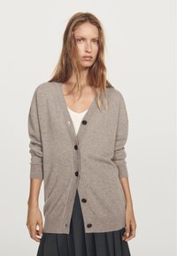 Grå cardigan av mjukt material med v-ringning, långa ärmar och mörka knappar fram. Bärs över en vit topp.