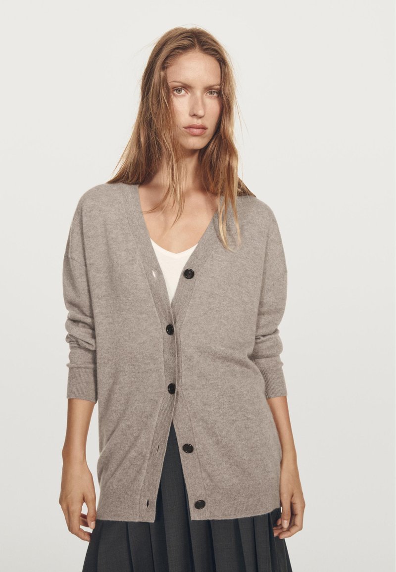 Grå cardigan av mjukt material med v-ringning, långa ärmar och mörka knappar fram. Bärs över en vit topp.