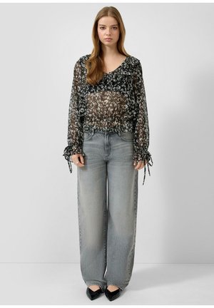 Junge Frau trägt eine durchsichtige schwarze Blumenbluse mit gerafften Manschetten, hoch taillierte, weit geschnittene, ausgewaschene graue Jeans und schwarze spitze Schuhe.