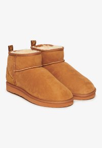 Botas de tobillo de gamuza color canela con un suave forro de pelaje en color crema. Presentan una costura decorativa y tiradores en la parte posterior. Suelas de goma planas.