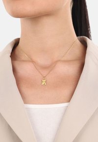 Collana con pendente a forma di orso in oro, indossata sopra un blazer beige chiaro, che mette in risalto la forma e la consistenza liscia dell'orso.