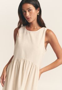 Beige ärmlös klänning med rund halsringning, rynkad midja och s flowing kjol. Tyget verkar texturerat och lätt.