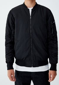 Veste bomber noire avec fermeture éclair à l'avant, poignets et ourlet côtelés, et poches latérales. Le tissu a une texture lisse avec un léger éclat.