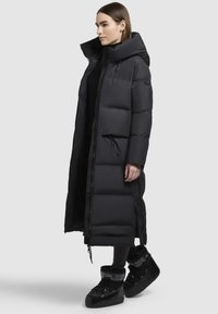 Garš melns pūkains jaka ar augstu apkakli, ar rāvējslēdzēju un auklu viduklim, ar lielām izšūtām daļām un gludu tekstūru.