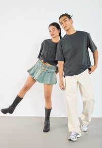 Two individuals pose, wearing oversized gray t-shirts. The female has a knot detail and a denim pleated skirt, paired with knee-high black boots. The male wears cream-colored pants and silver sneakers.