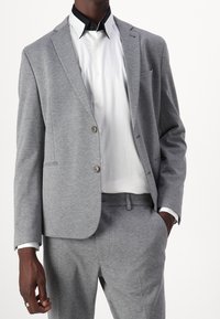 Szary garnitur o dopasowanym kroju z teksturowanym wykończeniem, z dwoma guzikami w marynarskim kroju i dopasowanymi spodniami. Biała koszula z czarnym kołnierzykiem.