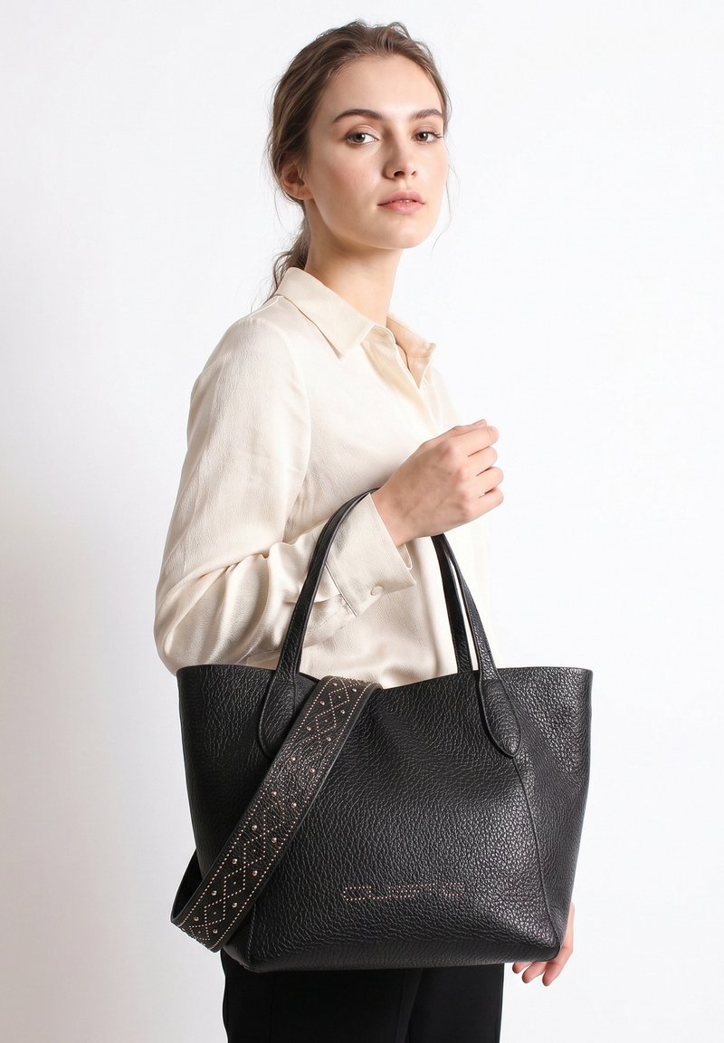 Jeune femme en chemise crème tenant un grand sac cabas en cuir noir texturé avec une bandoulière à motifs sur fond blanc.