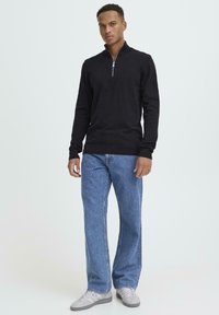 Juodas užtraukiamas megztinis iš megztos medžiagos, derinamas su šviesiai mėlynais plačiomis kelnėmis. Modelis dėvi pilkus sportbačius. Matomas tekstūruotos medžiagos detalė.