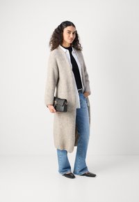 Light gray knitted cardigan with long sleeves, paired with a black top and white button-up shirt, flared blue jeans, and black flats.
