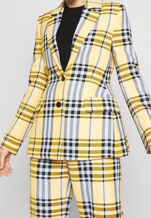 Żółta, czarna i jasnoniebieska kraciasta marynarka i pasujące spodnie noszone na czarnym prążkowanym topie, pokazane od szyi do połowy uda.