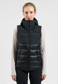 Schwarze isolierte Weste mit hohem Kragen, Reißverschluss und zwei Seitentaschen. Verfügt über eine puffy gesteppten Textur und minimales Logodesign.