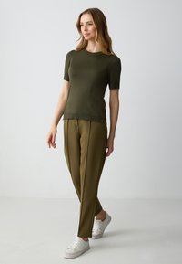 Olivgrüner kurzärmliger Pullover mit geripptem Ausschnitt, kombiniert mit olivgrünen, schmal geschnittenen Hosen und weißen Sneakers, vor einem schlichten Hintergrund.