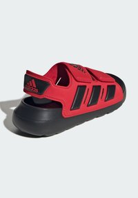 Sandale Adidas rouge et noire avec des lanières réglables en velcro, talon ouvert et semelle noire rembourrée sur un fond gris clair.