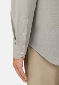 Camicia grigio chiaro con un motivo testurizzato, dotata di polsini su misura e bottoni beige. Accentata da un orlo leggermente curvo, abbinata a pantaloni beige.