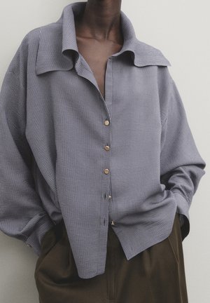 Person trägt ein locker sitzendes, graues Nadelstreifenhemd mit Knöpfen, das in eine hoch taillierte dunkelbraune Hose gesteckt ist, mit den Händen in den Taschen.