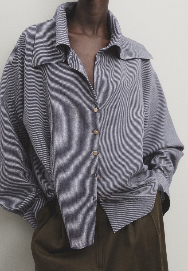 Personne portant une chemise ample grise à fines rayures boutonnée, rentrée dans un pantalon marron foncé taille haute, les mains dans les poches.