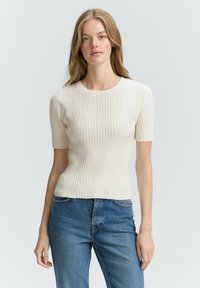 Crème ribgebreid knit top met korte mouwen en een fitted silhouet, met V-vormig detail aan de voorkant. Gesh Coupled met blauwe spijkerbroek.