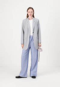 Grå kavaj med en enkel knapp, tillsammans med vida blårandiga byxor och en liten lavendelfärgad väska. Svarta skor kompletterar outfiten.