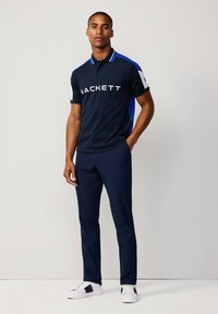 Mörkblå polo med vit logotyp "HACKETT," kontrasterande blå sidopaneler, i kombination med mörkblå byxor och vita sneakers.