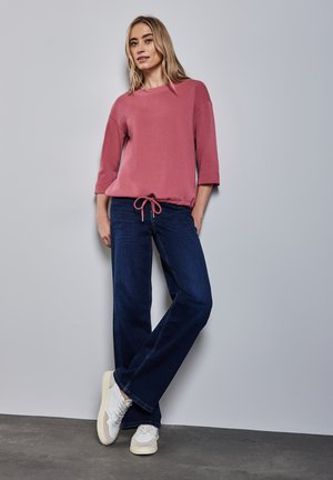 Light pink sweatshirt with a drawstring hem, paired with dark blue wide-leg jeans and white sneakers. Smooth texture, casual fit.