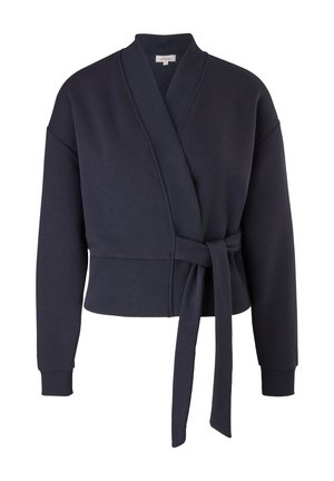 Kobieta ubrana w granatowy sweter z kopertowym zapięciem, zielone spodnie dresowe we wzory oraz jasnobeżowe sneakersy za kostkę, stoi na jednolitym tle.
