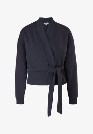 Kobieta ubrana w granatowy sweter z kopertowym zapięciem, zielone spodnie dresowe we wzory oraz jasnobeżowe sneakersy za kostkę, stoi na jednolitym tle.