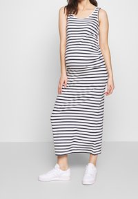 Schwangere Frau in einem ärmellosen Maxikleid mit schwarz-weißen Streifen und weißen Sneakers, die vor einem schlichten weißen Hintergrund steht.