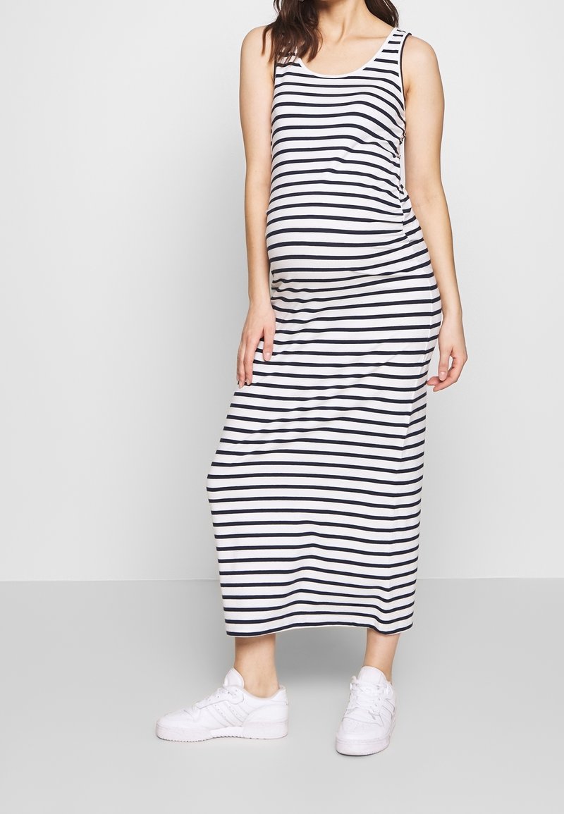 Schwangere Frau in einem ärmellosen Maxikleid mit schwarz-weißen Streifen und weißen Sneakers, die vor einem schlichten weißen Hintergrund steht.