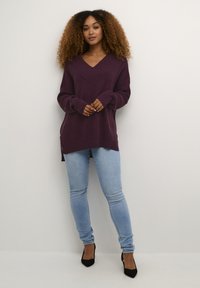 Overdimensioneret, ribbet lilla sweater med dyb V-udskæring, parret med lyseblå skinny jeans og sorte hæle. Tekstureret stof og sidelægninger synlige.