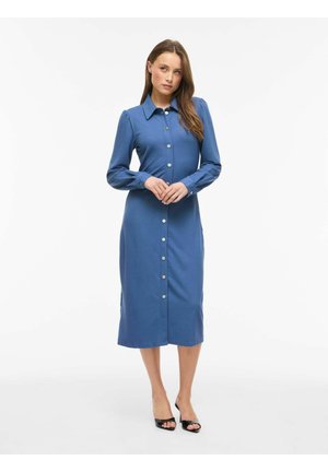 Kvinde står i en blå midi kjole med knapper og lange ærmer samt sorte peptoe-hæle mod en ensfarvet hvid baggrund.