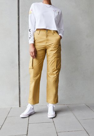 Personne portant un crop top blanc à manches longues avec des imprimés floraux, un pantalon cargo jaune moutarde et des baskets montantes blanches, debout à l'intérieur.