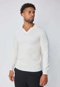 Pull en tricot blanc avec un col en V et un petit col, présentant des motifs côtelés verticaux et des manches longues. Texture lisse.