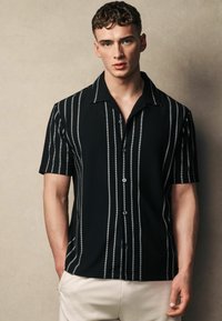 Camisa negra de manga corta con rayas verticales blancas, diseño de botones, textura acanalada y cuello con punta.