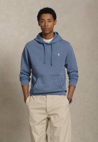 Lichtblauwe katoenen hoodie met een voorzak en een trekkoord, voorzien van een klein logo op de borst, gecombineerd met roomkleurige broeken.