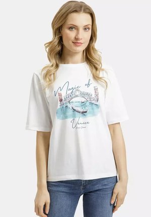 Młoda kobieta o długich blond włosach, ubrana w białą koszulkę z nadrukiem kanału w Wenecji oraz niebieskie dżinsy, stojąca na jednolitym tle.