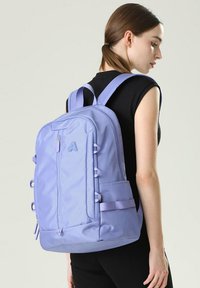 Sac à dos violet clair avec des bretelles rembourrées, plusieurs compartiments et un tissu texturé. Poche latérale avec détail de sangle ajustable.