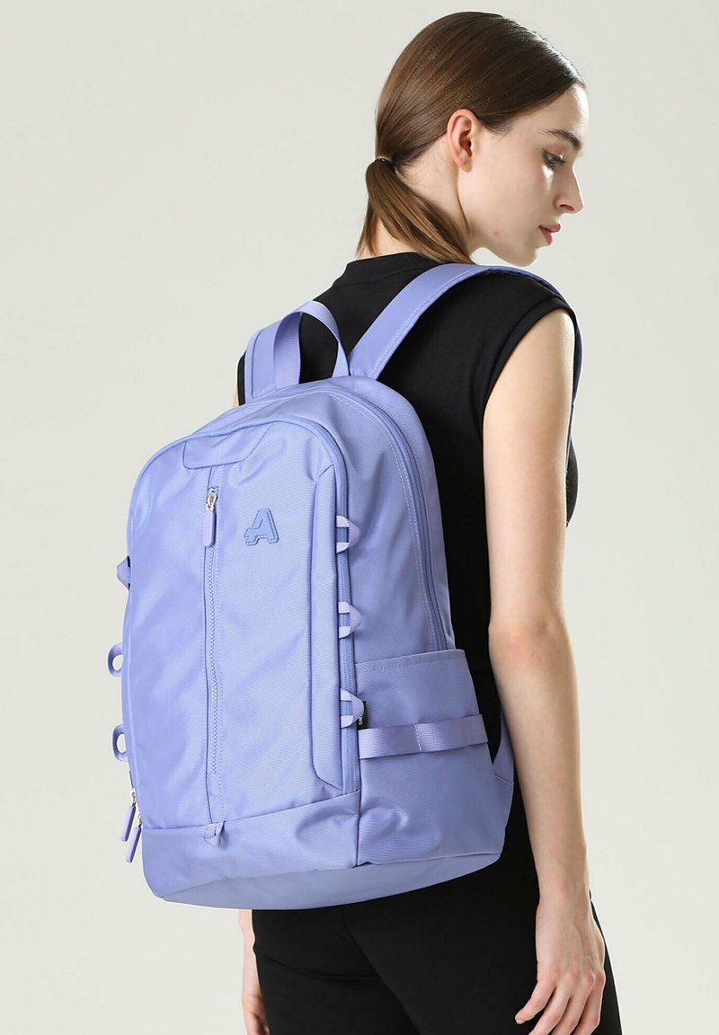 Sac à dos violet clair avec des bretelles rembourrées, plusieurs compartiments et un tissu texturé. Poche latérale avec détail de sangle ajustable.